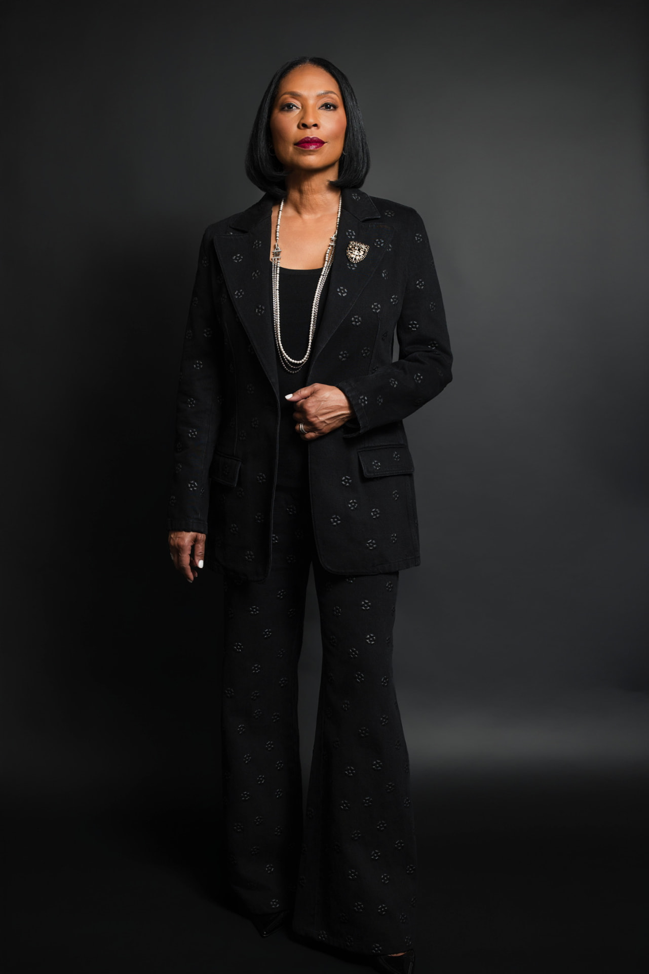 Patricia Lewis in a black Chanel suit with pearls, standing before a charcoal studio backdrop